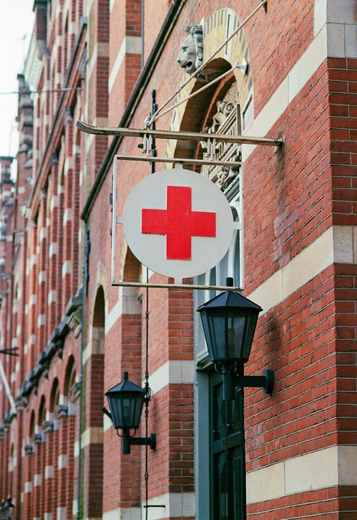Placa com o símbolo da Cruz Vermelha pendurada na fachada de um edifício antigo de tijolos vermelhos, sugerindo uma instalação médica ou hospitalar.