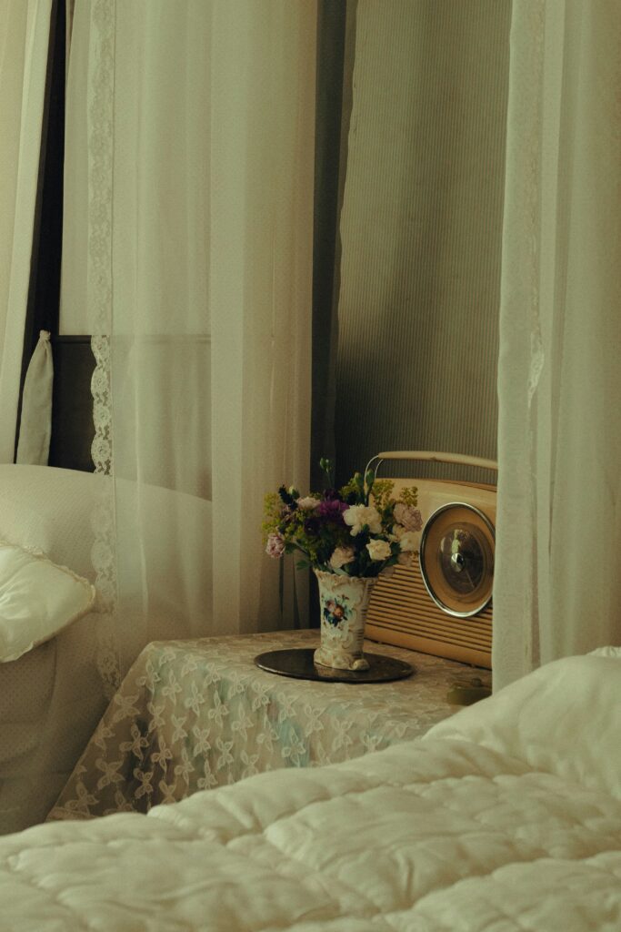 Quarto acolhedor de uma casa de repouso, com camas, cortinas claras, mesa lateral decorada com flores e rádio.