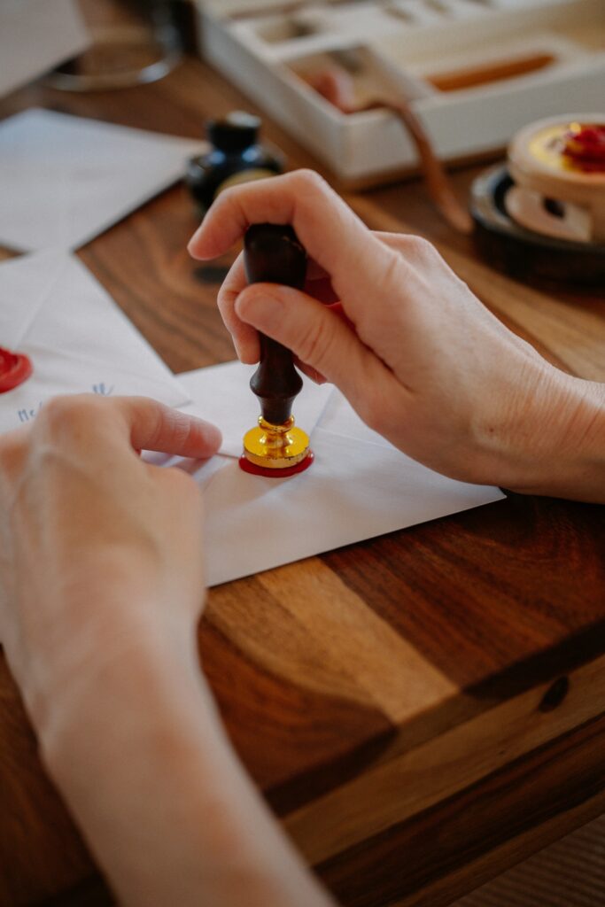Mãos usando um carimbo de latão para aplicar lacre vermelho sobre um envelope branco em uma mesa de madeira, remetendo à autenticação de documentos e à Apostila de Haia.